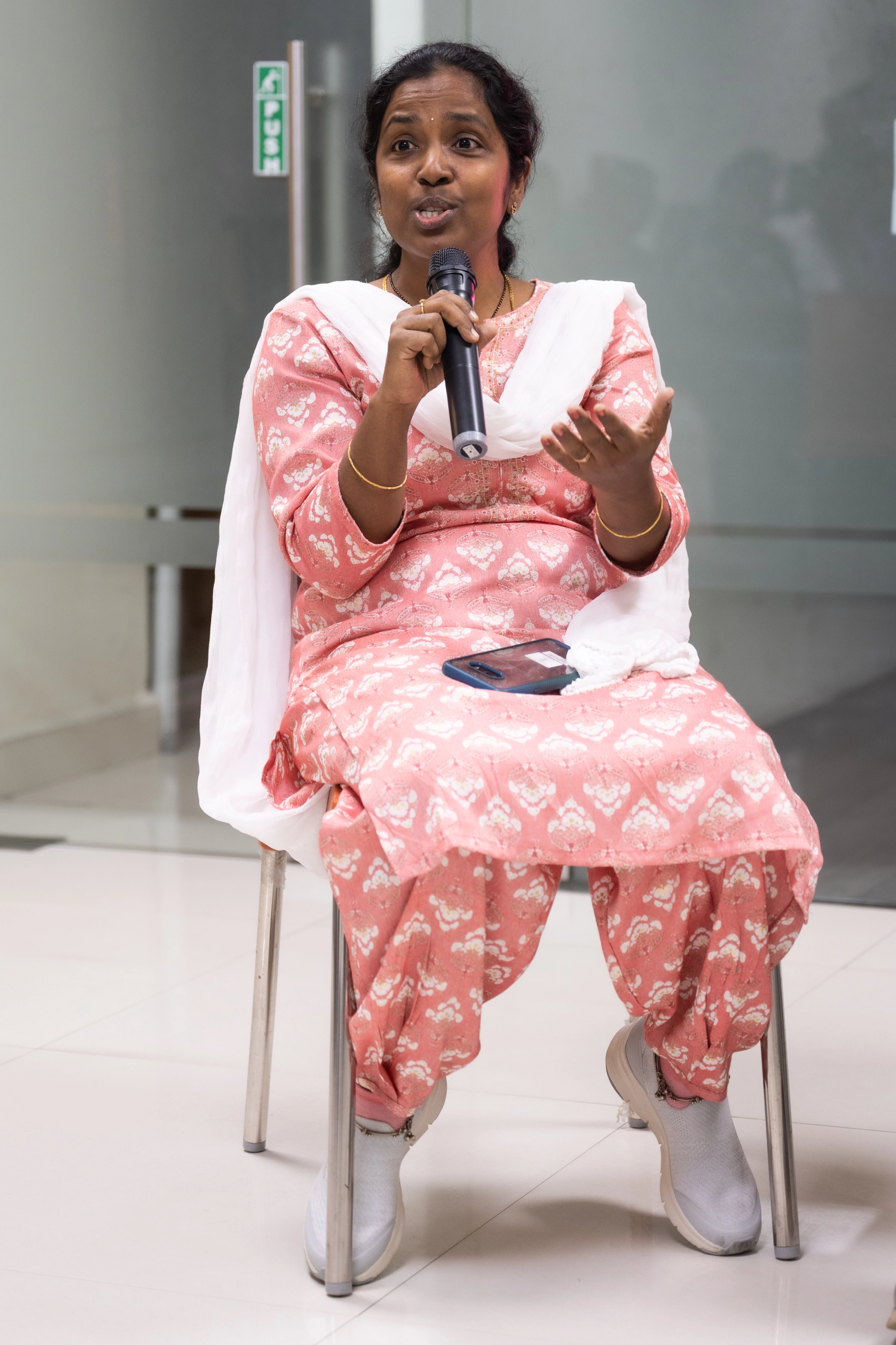 Woman in pink patterned traditional attire speaking into a microphone while seated on a metal chair indoors.