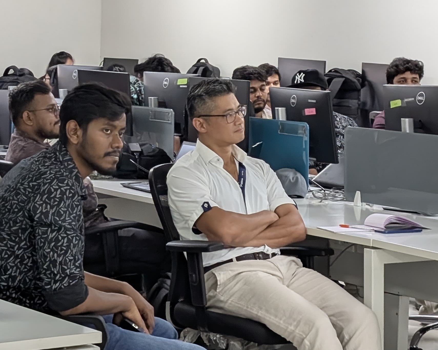 Group of people sitting attentively in an office with multiple computer monitors on desks.