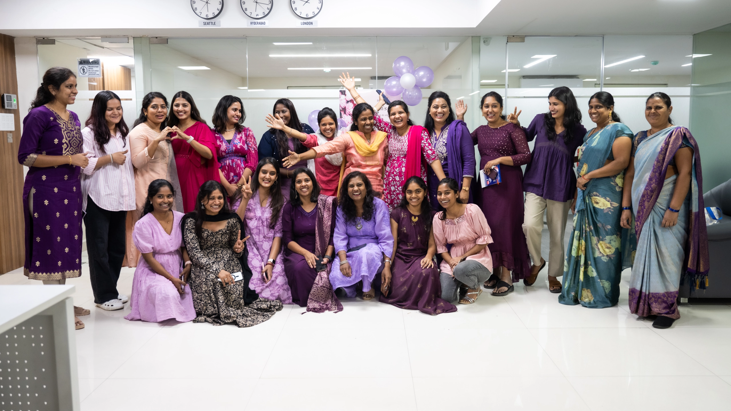 Group of smiling women dressed mostly in purple and pink, posing indoors with some kneeling and others standing.