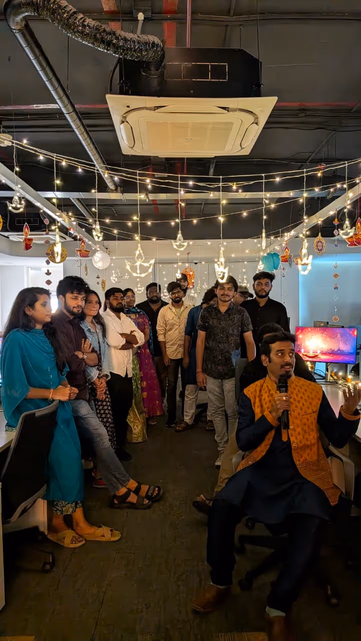 Group of people standing and sitting in a decorated office space with hanging lights and traditional ornaments, one man speaking into a microphone.