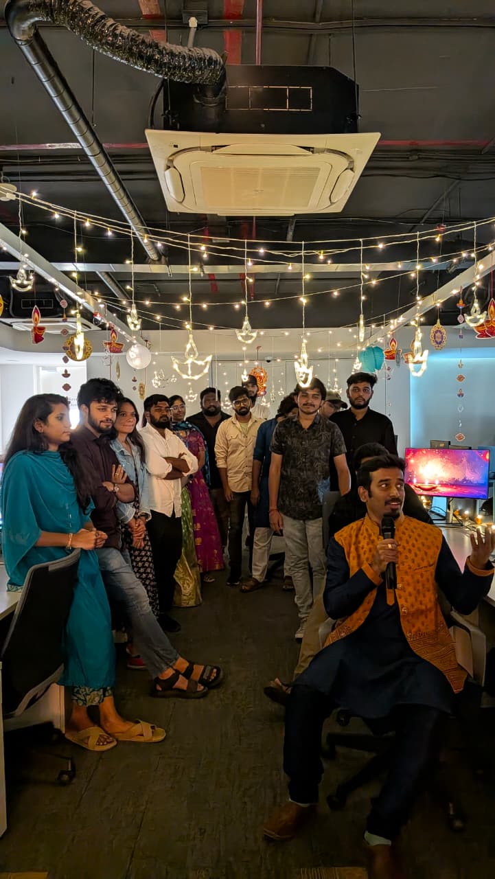 Group of people standing and sitting in a decorated office space with hanging lights and traditional ornaments, one man speaking into a microphone.