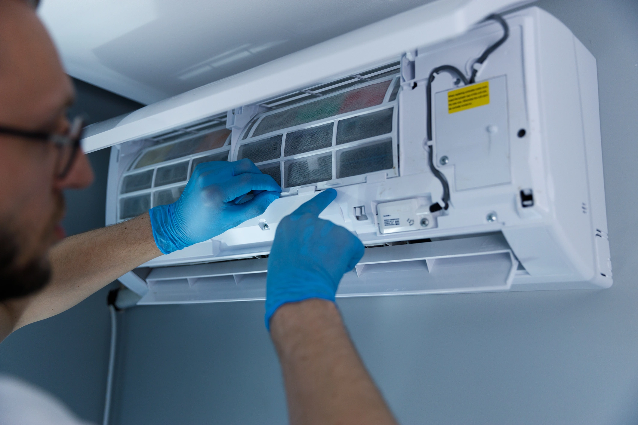 A person wearing blue gloves is cleaning or repairing a wall-mounted air conditioning unit, focusing on the filter. 