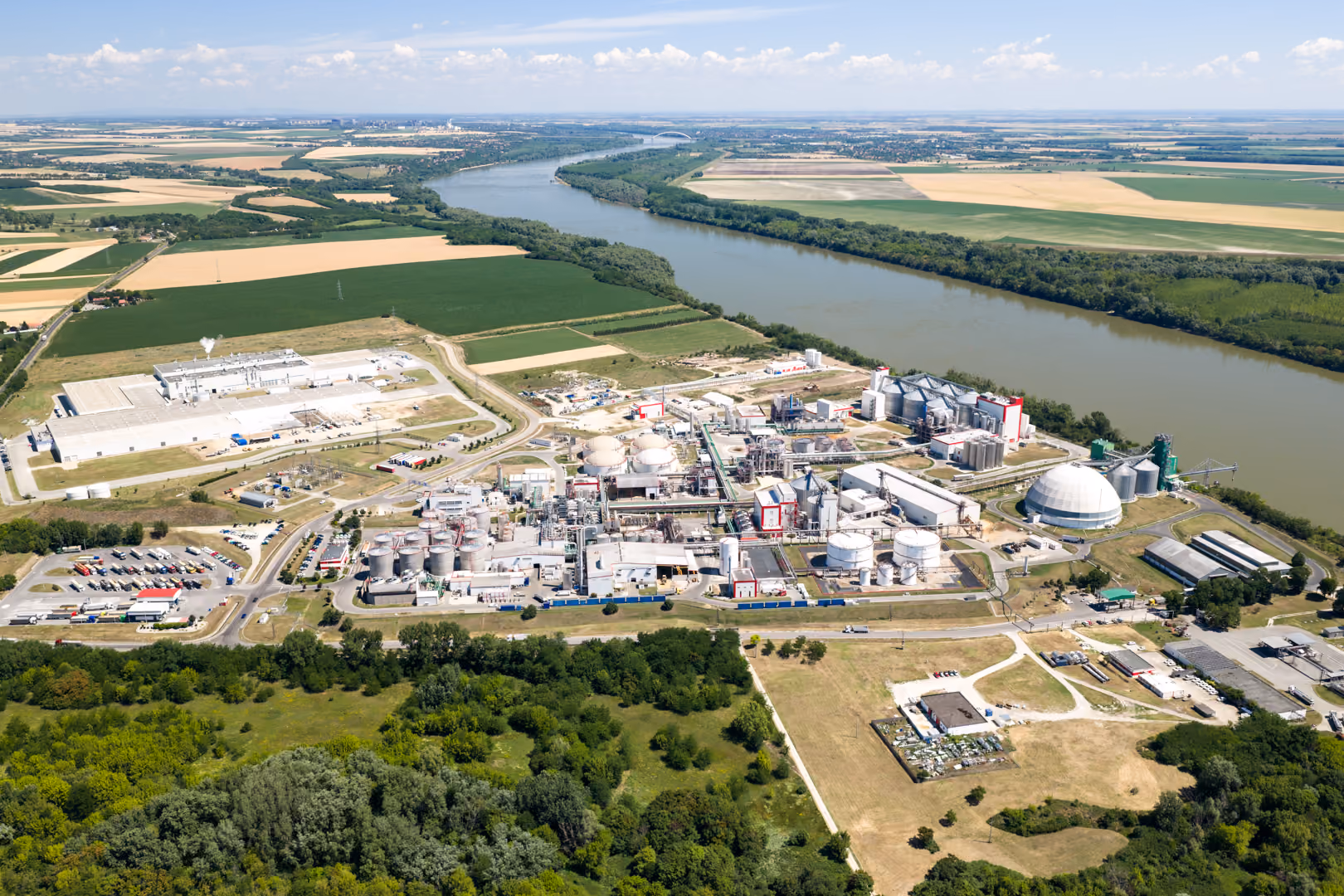 Aerial view of an industrial refinery complex with multiple storage tanks, silos, and buildings situated next to a wide river, surrounded by green fields and patches of forest under a partly cloudy sky.