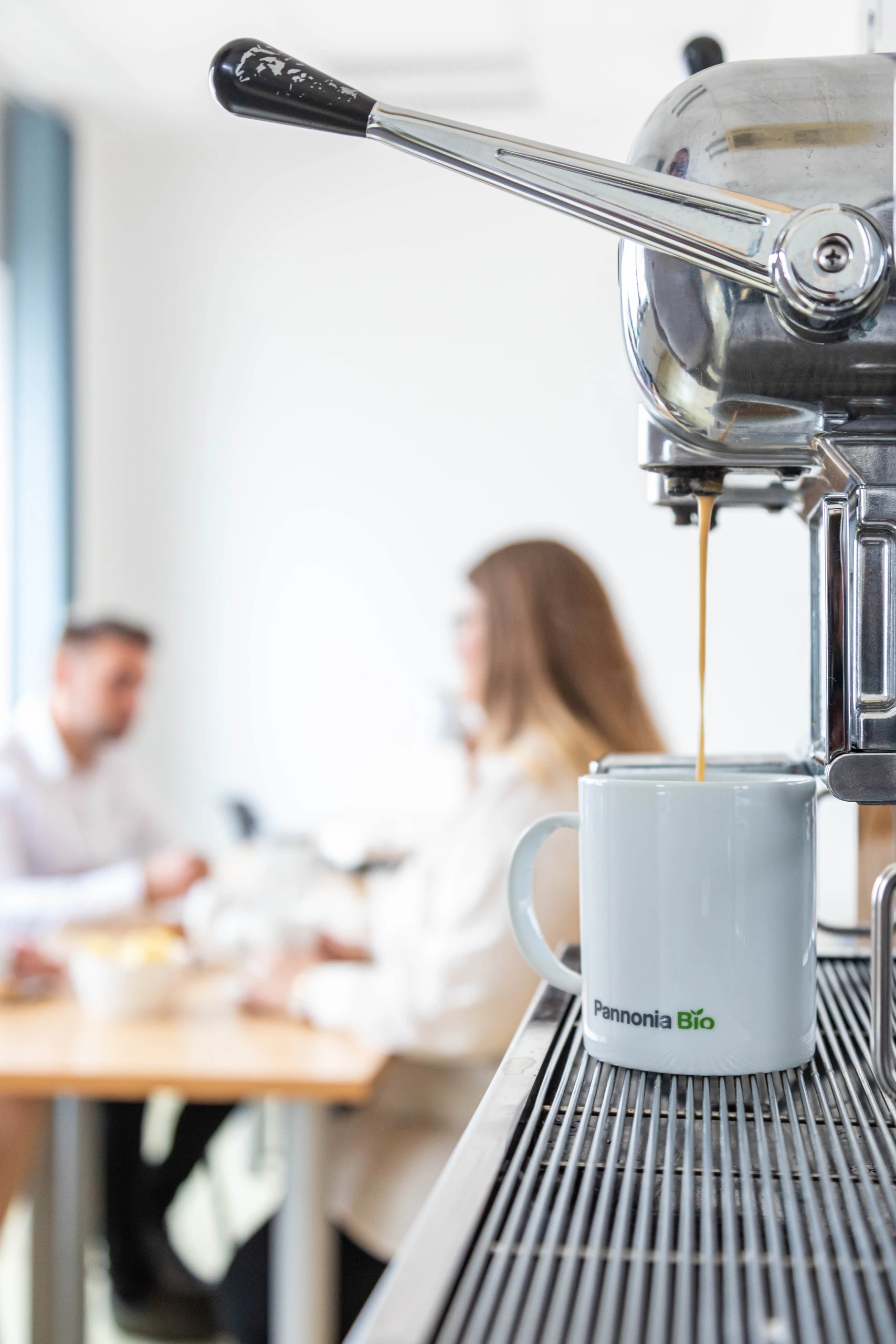 Coffee pouring into a Pannonia Bio mug, with colleagues chatting in the background