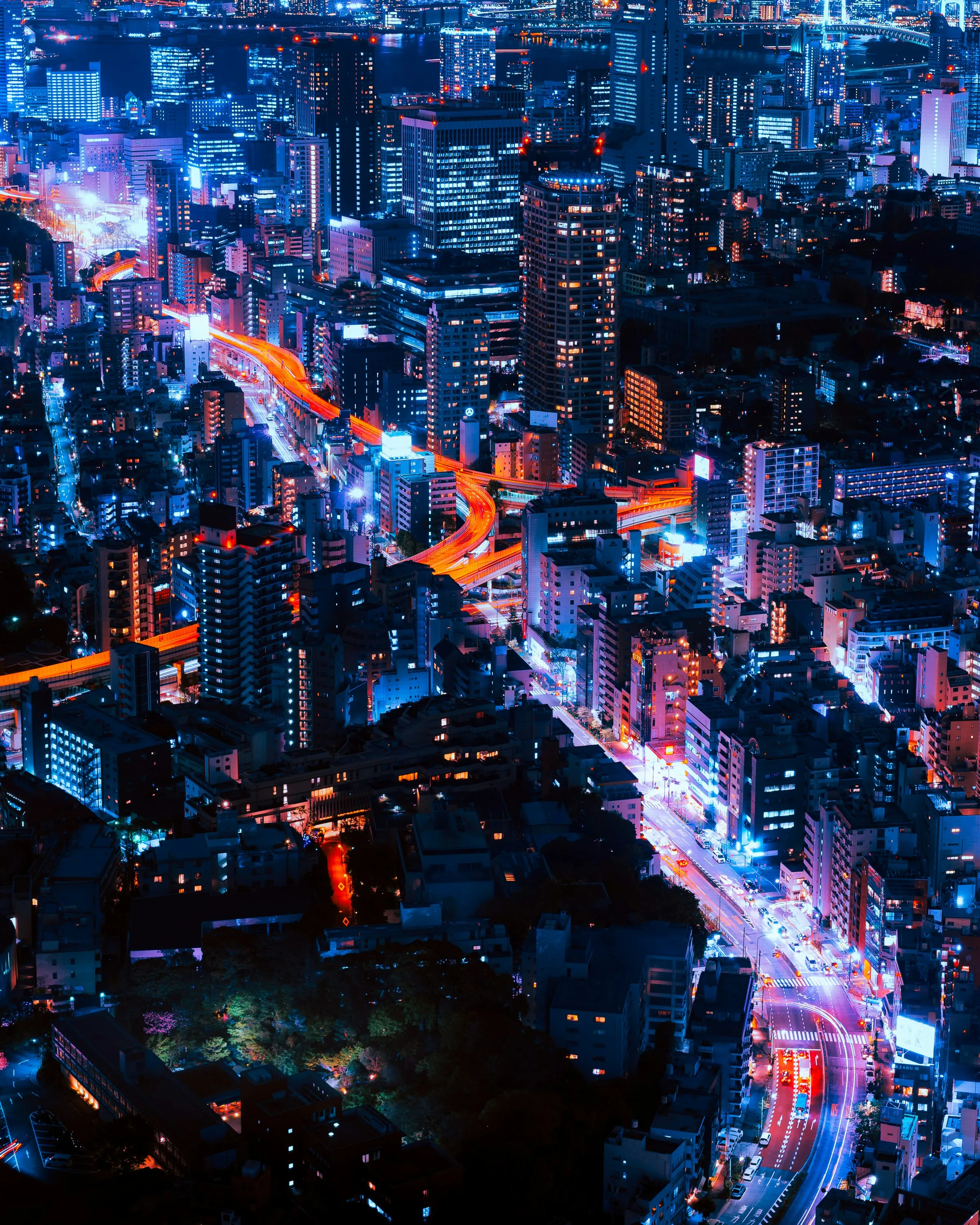 Aerial nighttime view of a cityscape with densely packed high-rise buildings illuminated in blue and white lights, featuring winding highways glowing in bright orange-red that curve through the urban area.