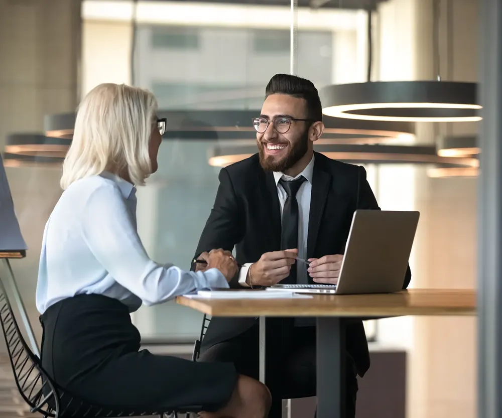Two business professionals, a man in a suit and glasses and a woman with blonde hair and glasses, smiling and talking across a table with a laptop in an office setting.