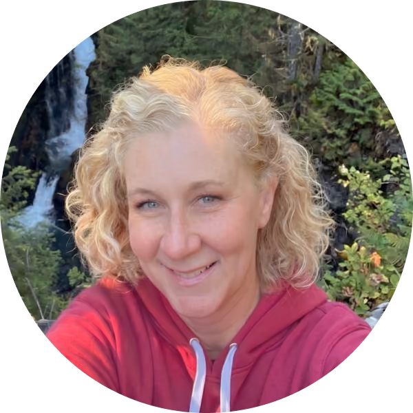 Smiling woman with curly blonde hair wearing a red hoodie with a waterfall and greenery behind her.