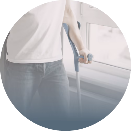Person using a gray crutch near a window, wearing a white shirt and blue jeans.