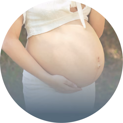 Close-up of a pregnant woman’s bare belly with her hands gently cradling it, wearing a white top and white pants.