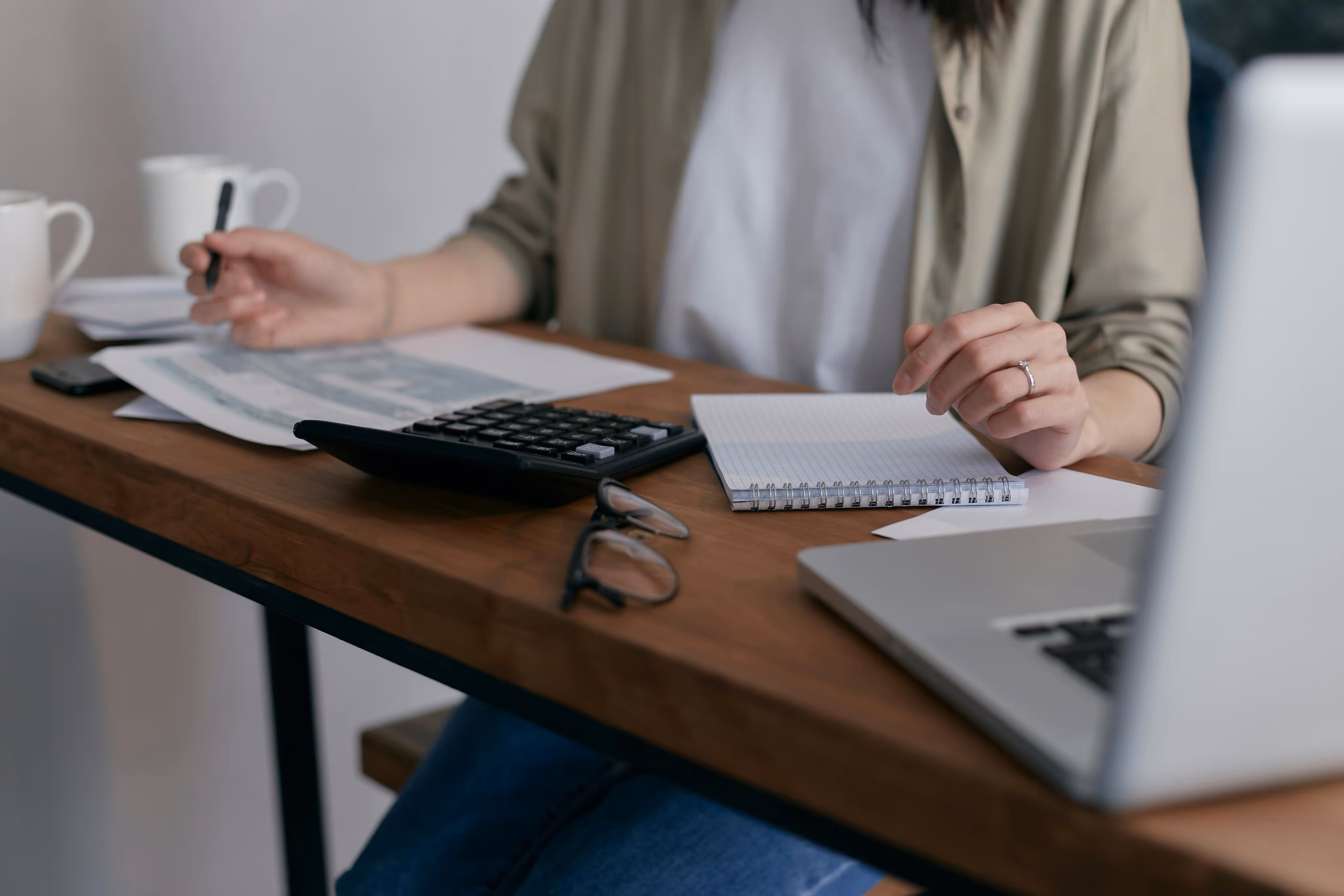 Person working at a wooden desk with a laptop, calculator, notebook, papers, and glasses.