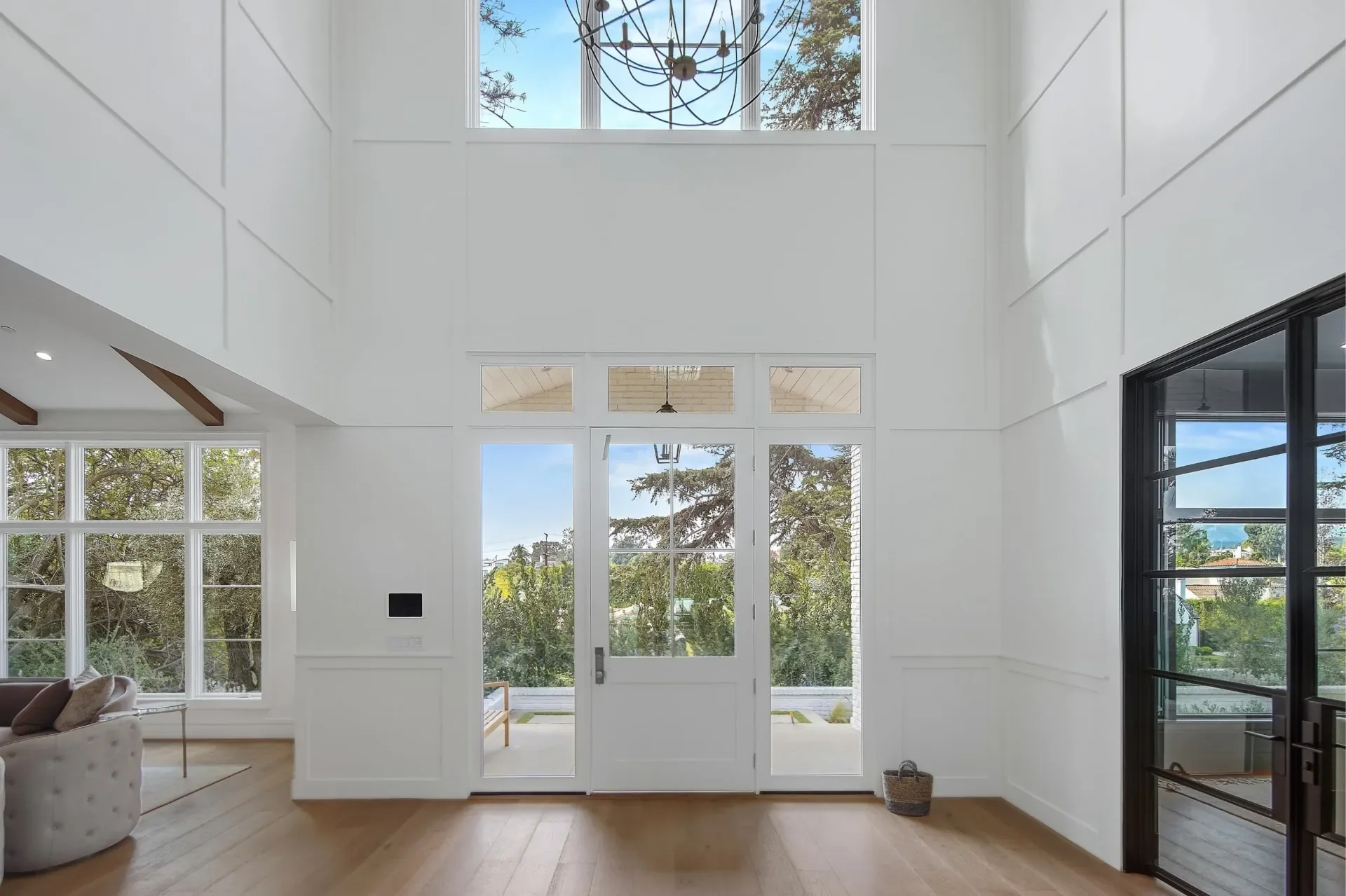 Double-height South Bundy entry with white wall panels, tall dark-framed side glazing and a chandelier above the front doors.