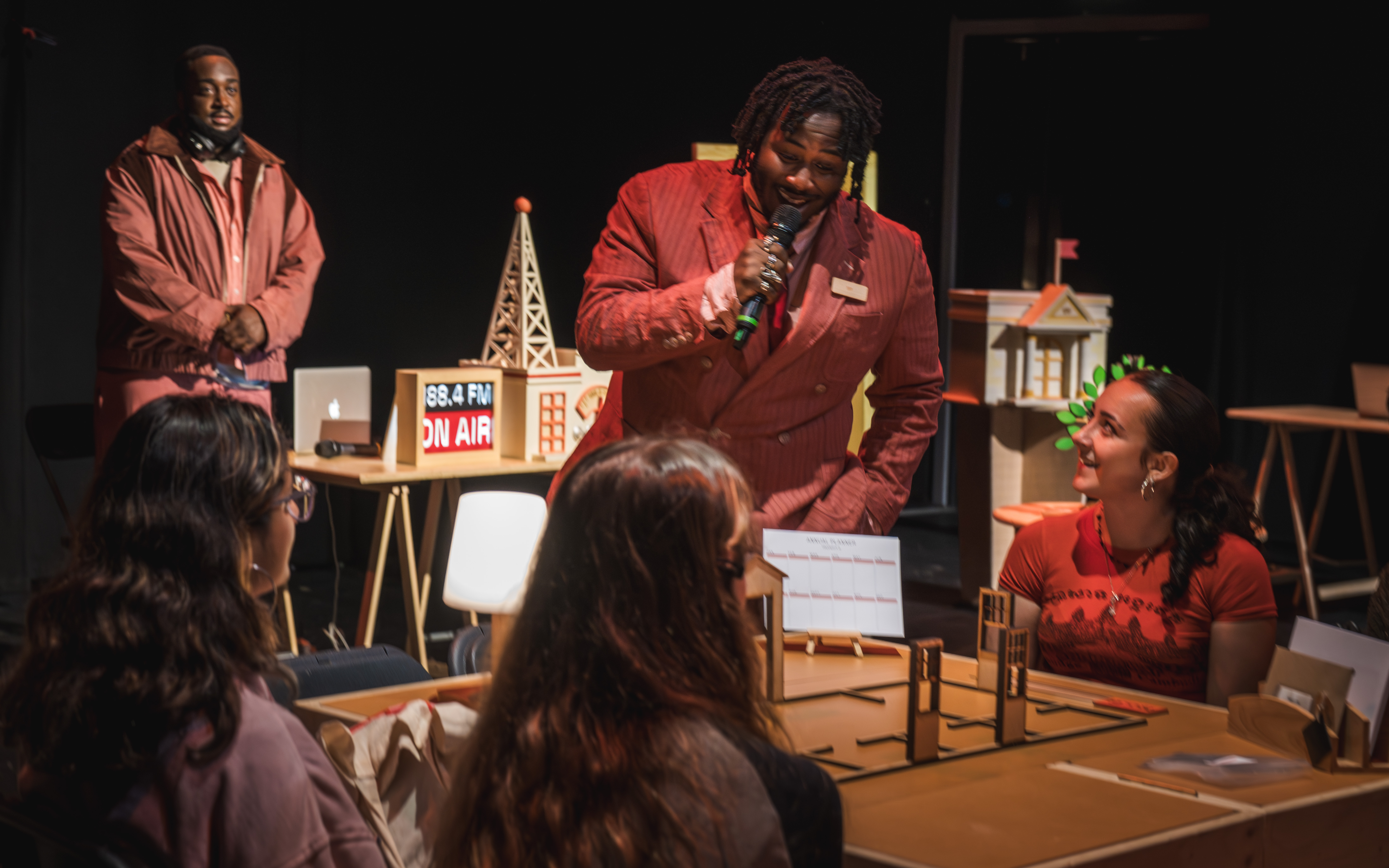 One man dressed in red holding a microphone addresses a group of audience members sitting around a wooden floor plan
