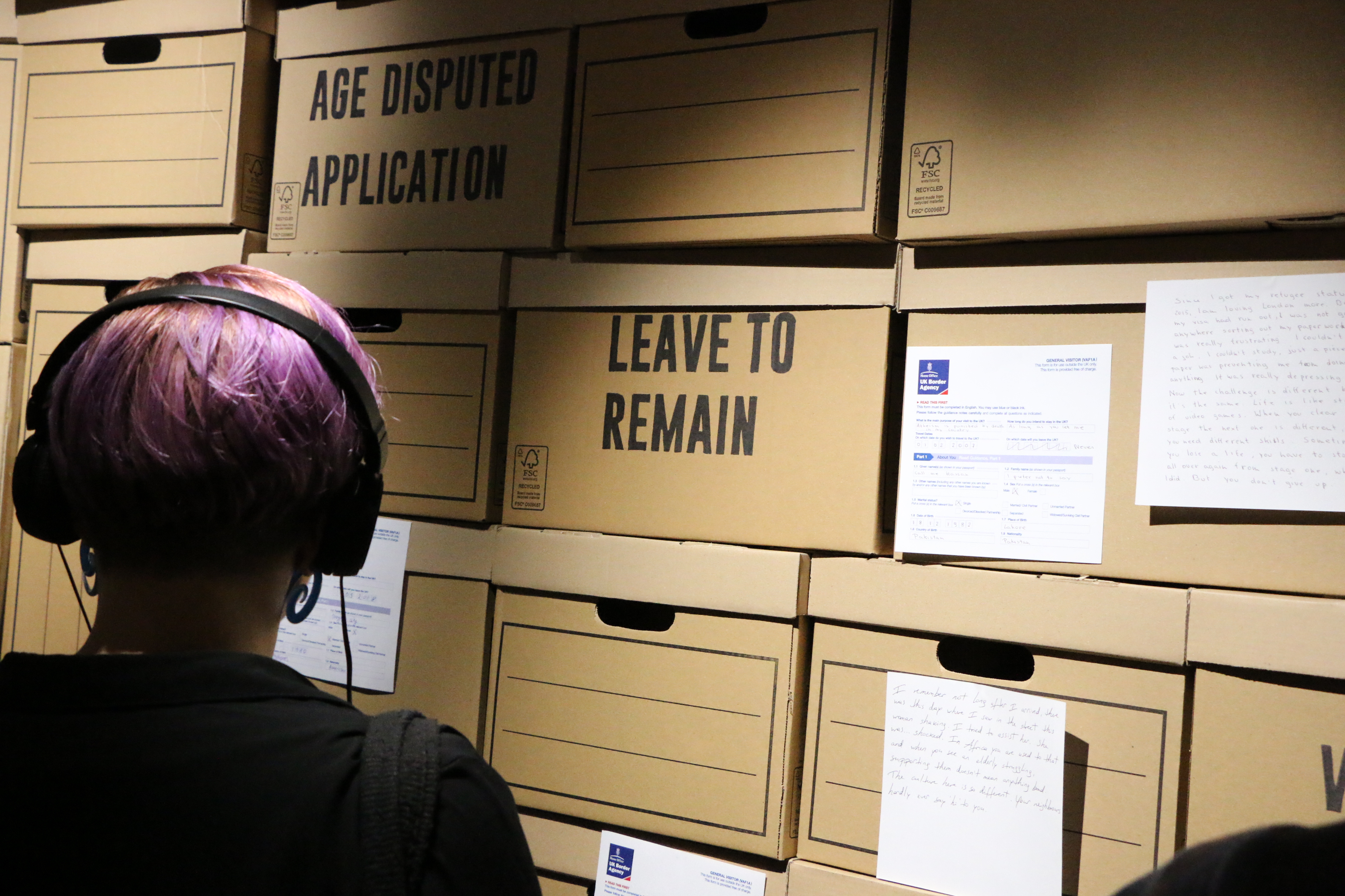 Audience member wearing headphones looking at archive boxes marked "Leave to remain"