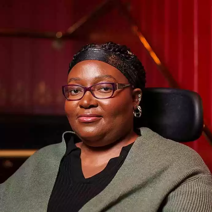 Matilda is a Black person who sits facing the camera, with a slight smile, against a red backdrop, wearing glasses, a dark top, and a green cardigan. Their braids are styled back with a headband, and they wear small hoop earrings. A wheelchair headrest is visible behind them.