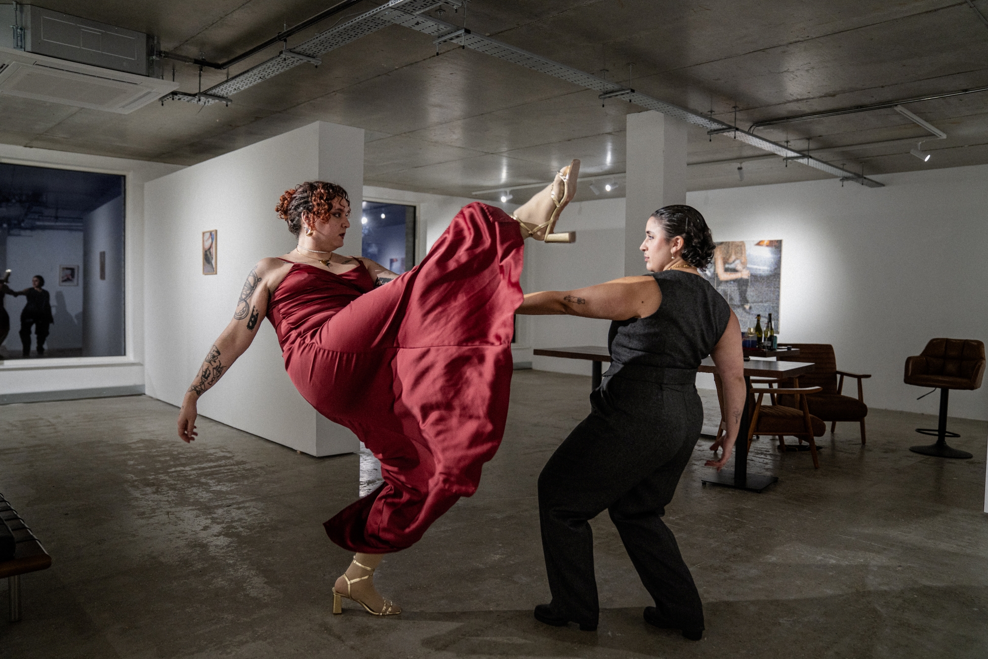Two chunky people in front of an art gallery background are looking at each other holding hands. One of them is a white transfem person with curly red hair and a red satin dress, they have one of their legs mid high kick and the satin dress is floating under it. The other one is a non-binary masc person with olive skin wearing dark grey waistcoat and trousers and their short curly hair is slicked back
