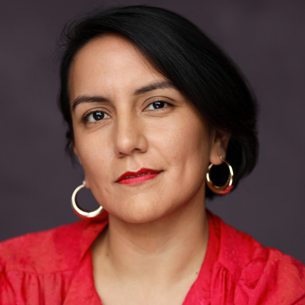 A headshot of Pepa, she has short, dark hair tucked behind her ears. She’s looking directly at the camera with a calm, subtle smile. She wears large gold hoop earrings, bold red lipstick and a vibrant pink button-up blouse with a delicate, textured pattern. The background is a solid dark charcoal grey.