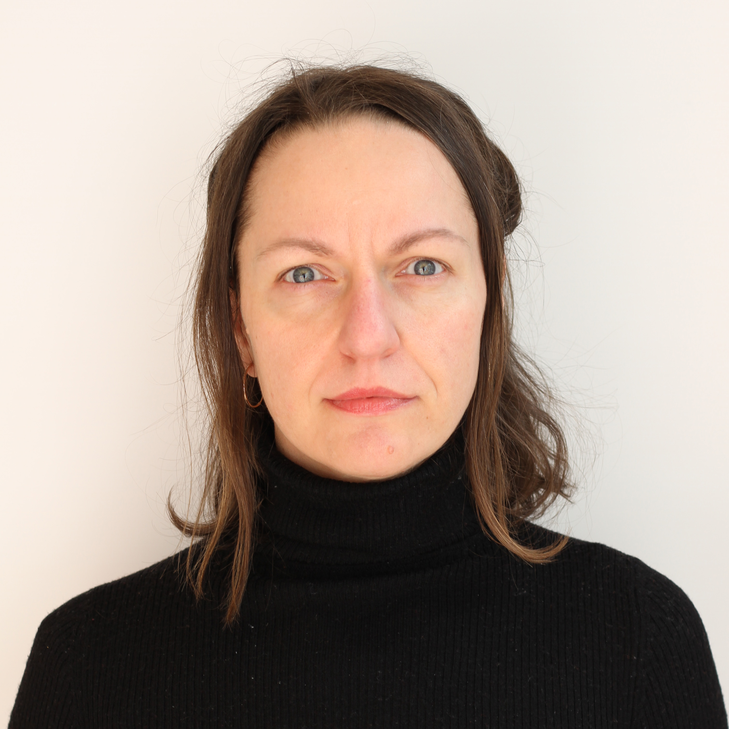 A medium close-up, front-facing portrait of a woman - Margot - standing against a plain, off-white wall. They have light-colored eyes and brown, shoulder-length hair that is tucked behind their ears, with a few loose strands framing their face. The person is wearing a black, ribbed turtleneck sweater and a small gold hoop earring is visible in their right ear. They have a neutral, direct expression, looking straight into the camera. The lighting is bright and even, casting a soft shadow against the wall behind them.