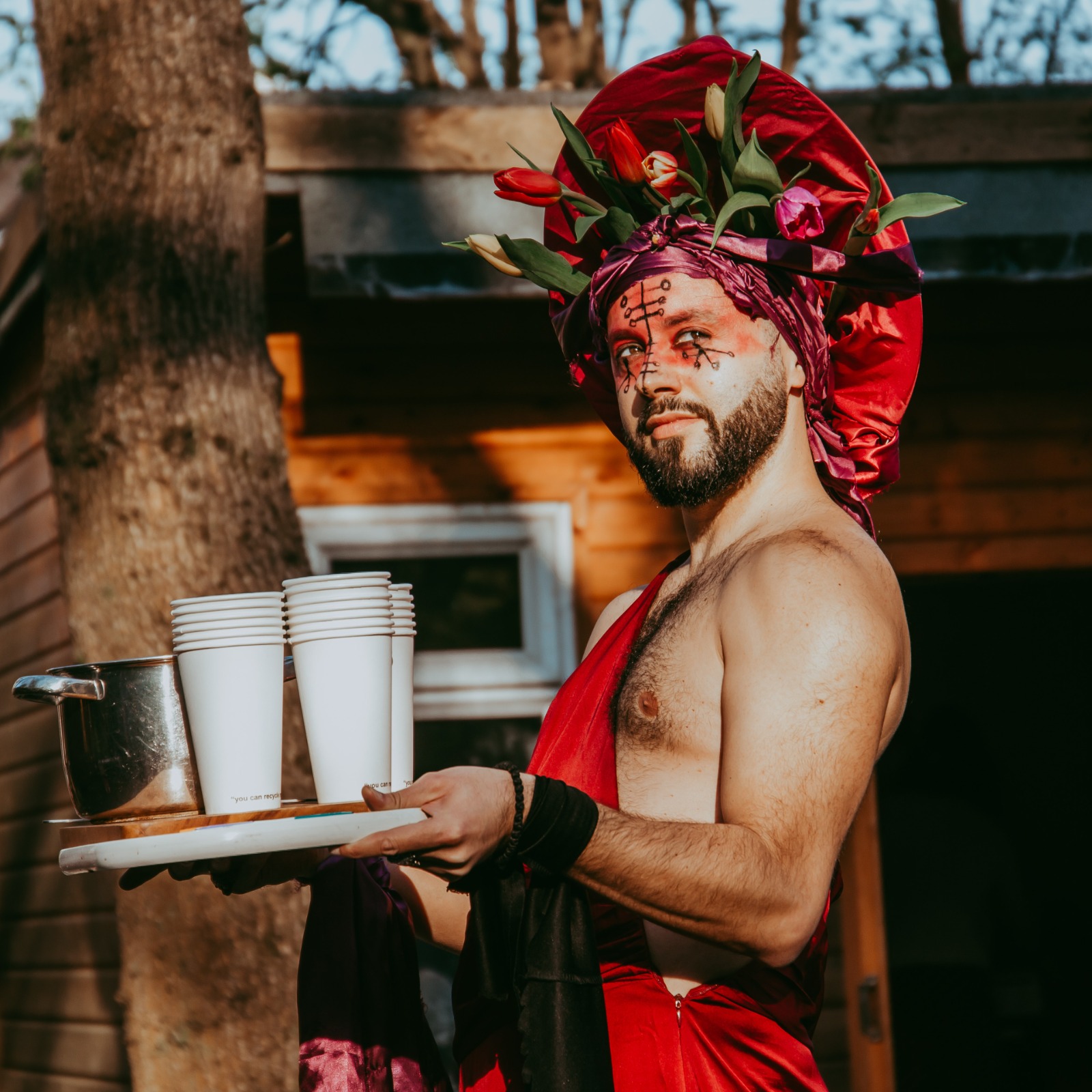 A performer wearing makeup and a colourful flower headpiece carrying a plate with a coffee pot and cups