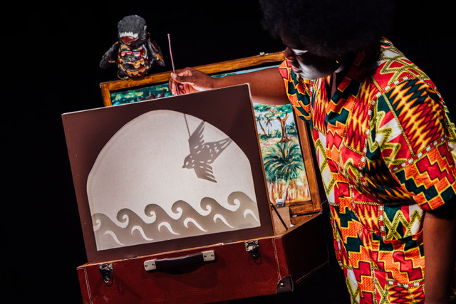 A performer in a colourful African top creating a bird shadow out of a suitcase