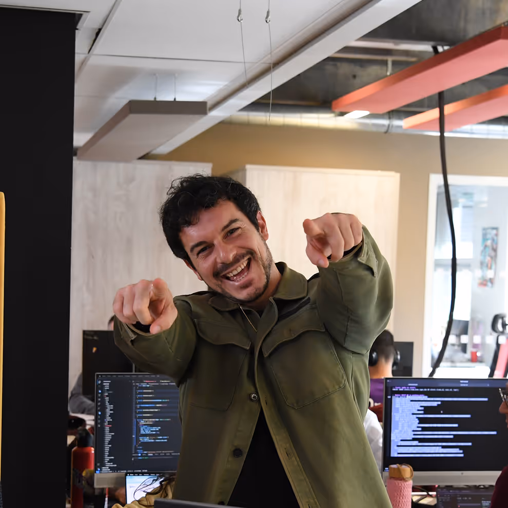 Smiling man in a green jacket pointing towards the camera in an office with computer screens in the background.