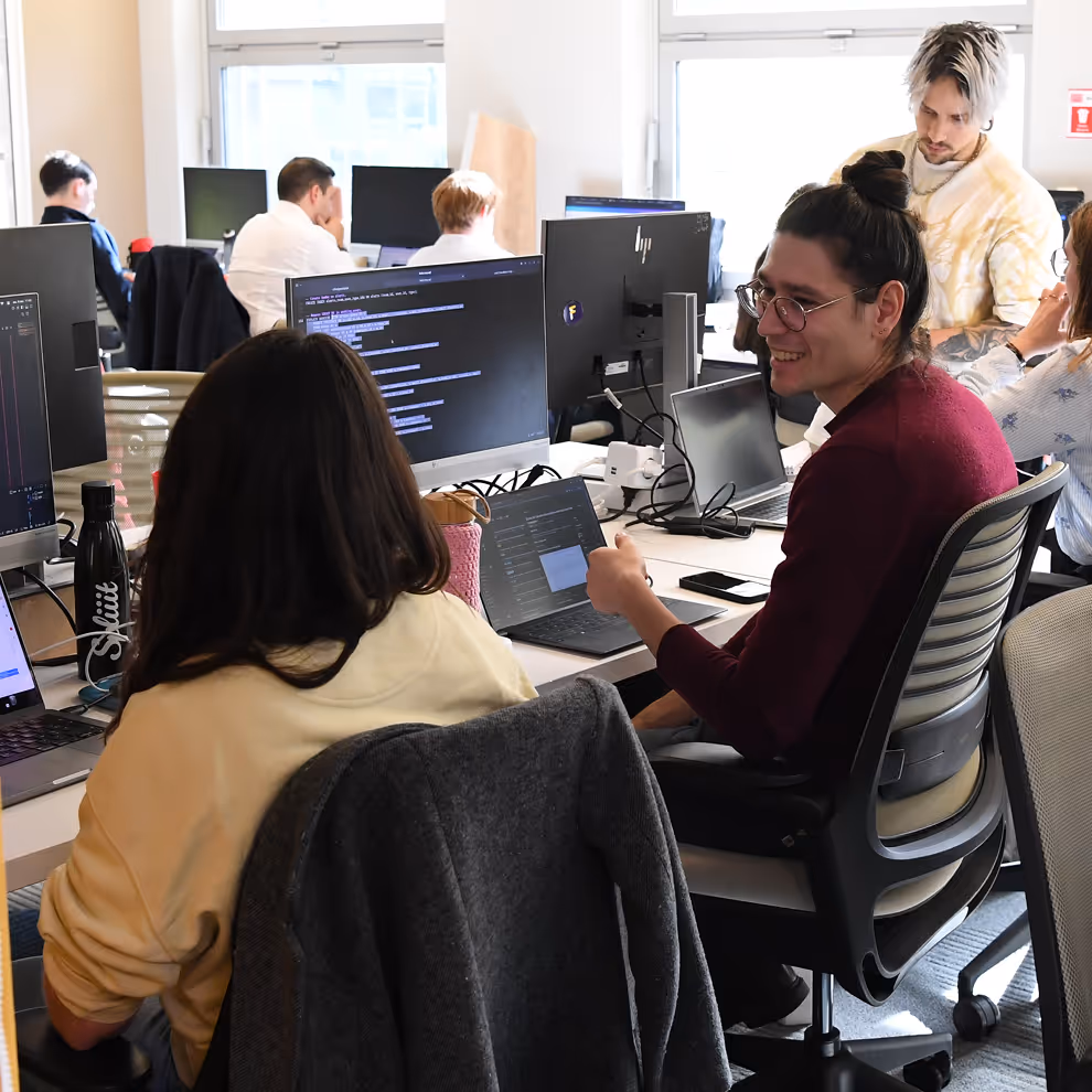 Several people working together at computers in a bright office, one person smiling and talking to a colleague.