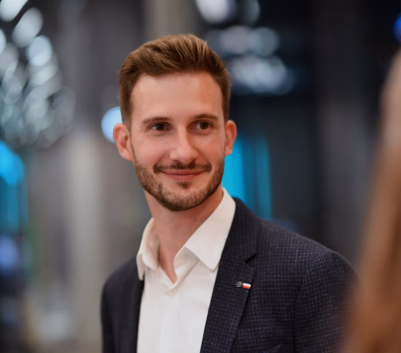 Smiling man with short brown hair and beard wearing a dark blazer and white shirt in a blurred indoor setting.