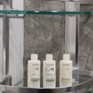 Three white bottles of Nutrei Amazonia balancing shampoo displayed on a glass shelf with a gray textured background.