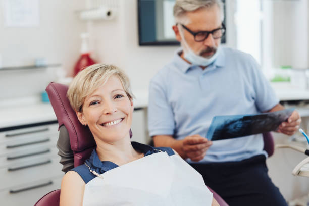 Smiling friendly woman patient in a dental surgery Smiling friendly woman patient in a dental surgery looking at camera as the senior dentist checks her X-rays Tasteful, anonymized portrait of an adult patient post-visit, seated and relaxed, soft smile; dentist in background reviewing images. stock pictures, royalty-free photos & images