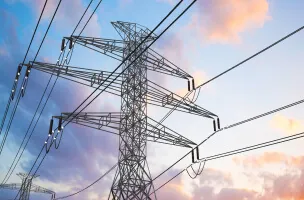 Uma torre de transmissão de energia elétrica de metal contra um céu com nuvens coloridas pelo pôr do sol, destacando a infraestrutura de energia elétrica.