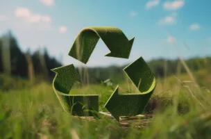 Símbolo de reciclagem composto por três setas verdes entrelaçadas em formato triangular, feitas de material que parece folhas, posicionado sobre a grama, representando a sustentabilidade e reciclagem na natureza.