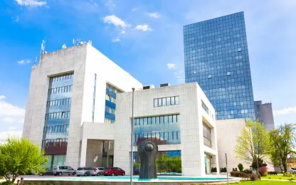 Edifício moderno de vários andares com fachada de concreto e vidro sob um céu azul claro, com vegetação em frente e uma escultura artística ao lado da entrada.