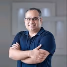 Smiling man with glasses and crossed arms wearing a navy blue polo shirt in a blurred indoor setting.