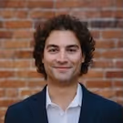 Smiling man with curly dark hair wearing a white shirt and dark blazer in front of a brick wall.