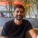 Smiling man with a beard and short hair sitting indoors with plants and a window in the background.
