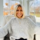 Smiling woman with blonde hair wearing a cozy light gray sweater seated indoors near large windows with autumn foliage outside.