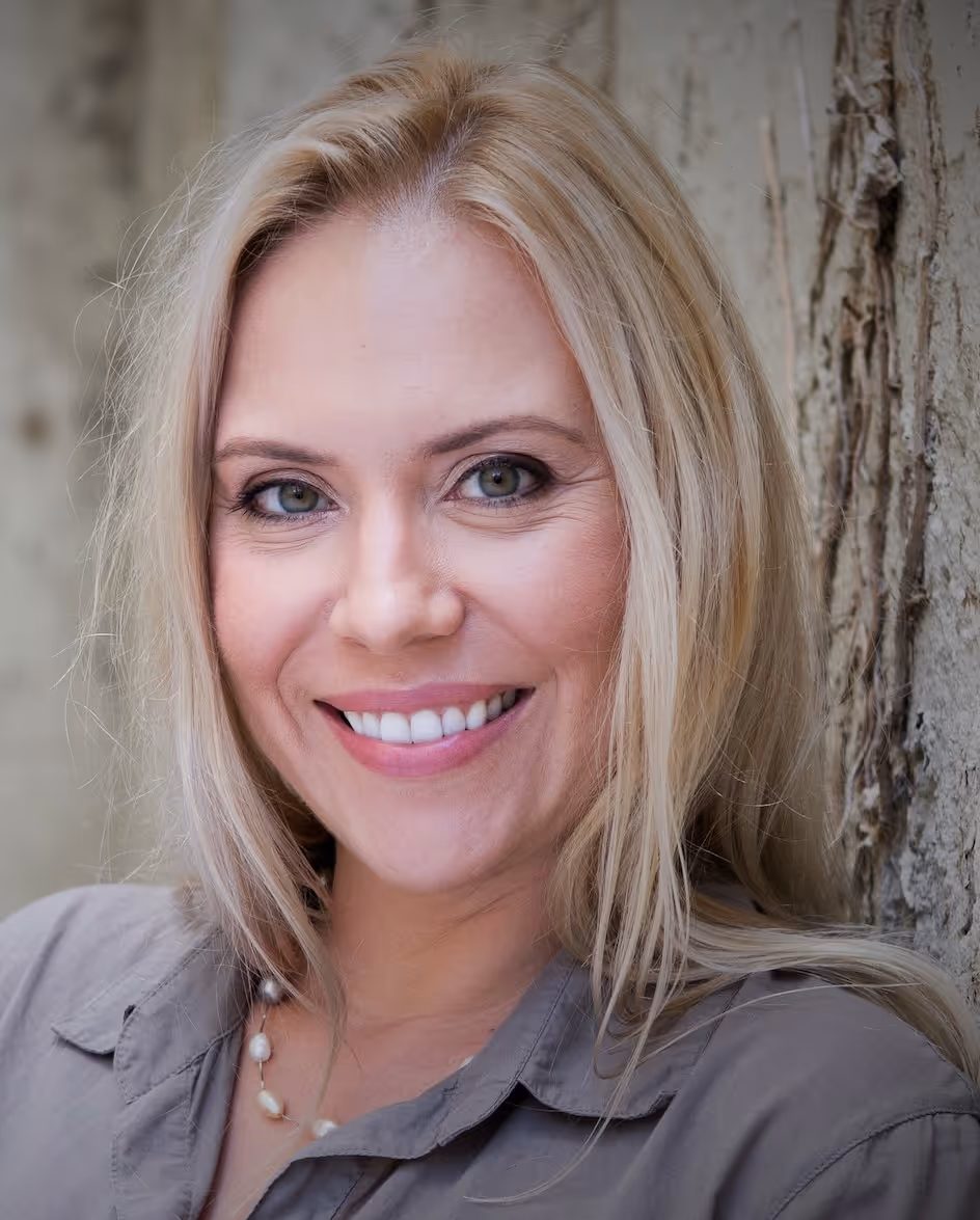 Close-up of a smiling woman with blonde hair wearing a gray shirt and a pearl necklace, leaning against a textured wall.