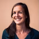 Smiling woman with long dark hair wearing a dark green top against a plain peach background.