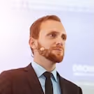 Man with a beard and mustache wearing a suit and tie, speaking with a headset microphone.