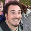 Man with short dark hair and goatee smiling, wearing a green shirt and black backpack, with a brown and white dog on leash and a baby in a blue outfit in the background.