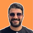Smiling man with dark hair, beard, and sunglasses against an orange background.
