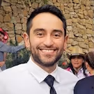 Smiling man with dark hair and beard wearing a white shirt and black tie, standing in front of a stone wall with people in the background.