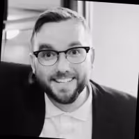 Smiling man with glasses, beard, and short hair wearing a suit jacket and white shirt.