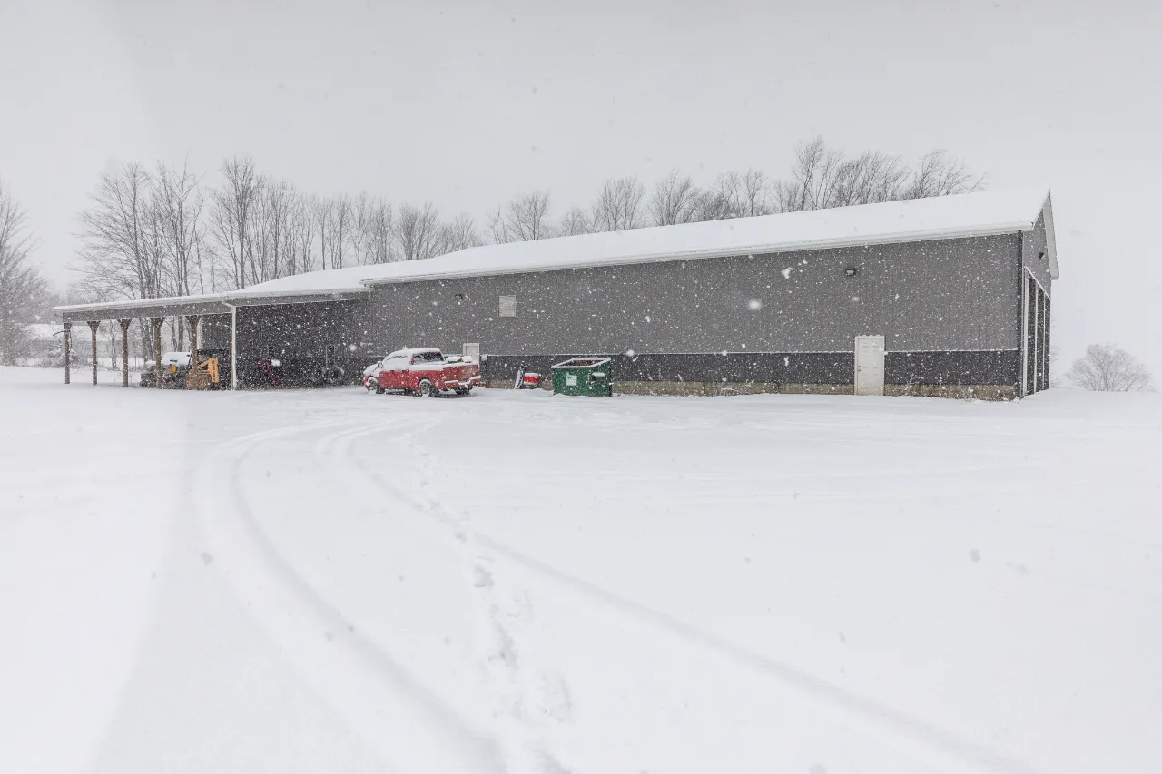 Heavy-duty repair shop exterior during snowfall with a red pickup and equipment parked under the awning.