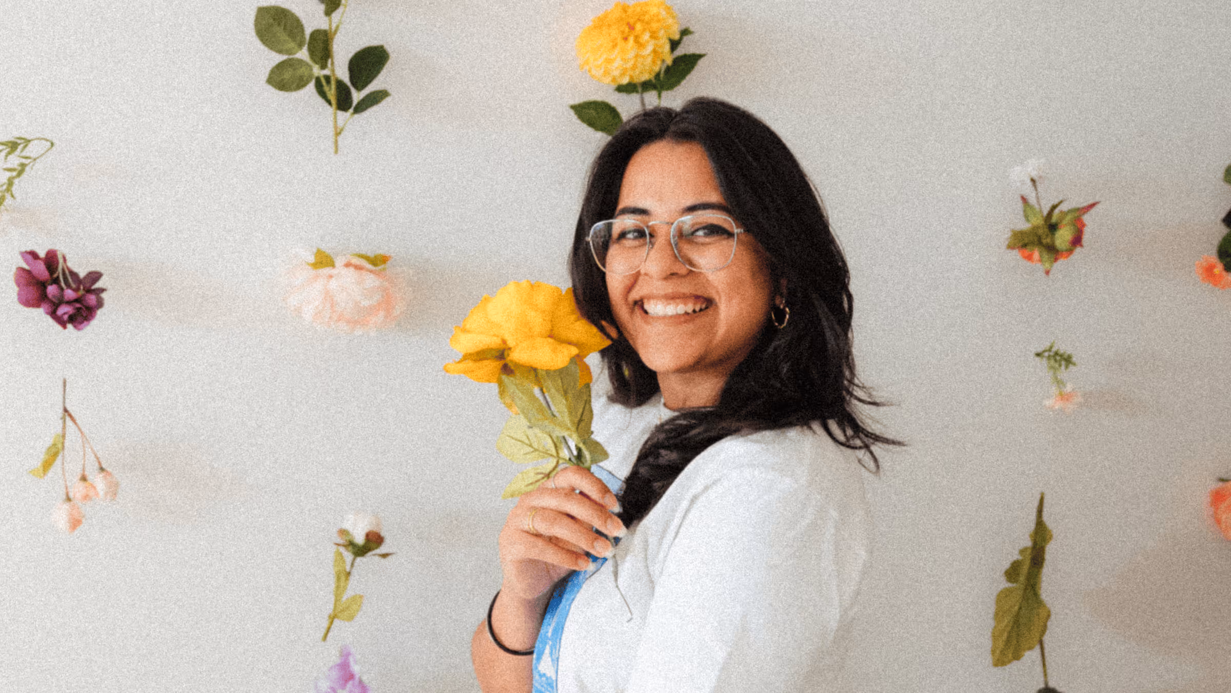 Smiling woman wearing glasses holding a yellow flower with various flowers hanging on a light background.
