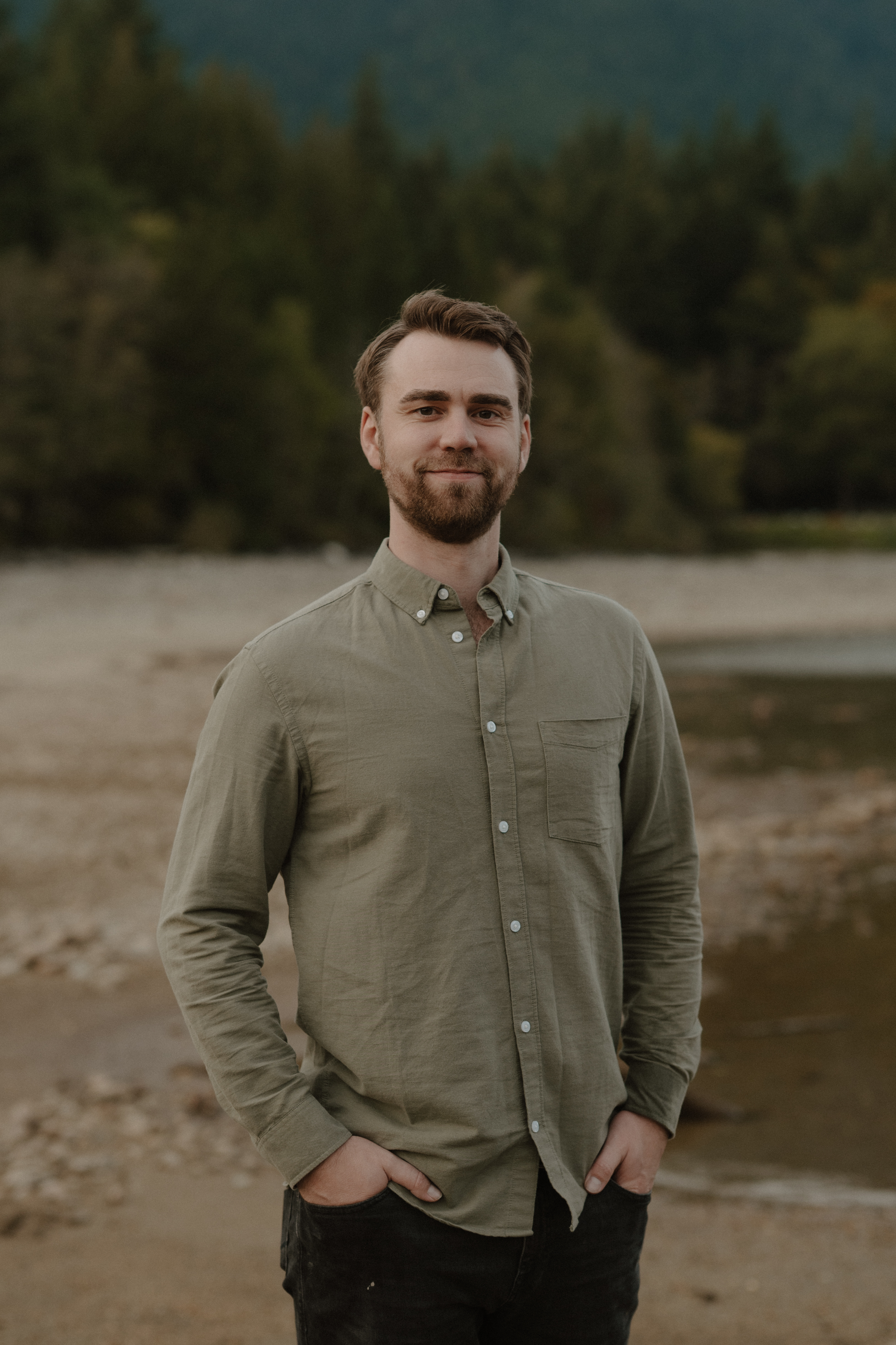 Portrait of Chris Aitken, a young individual with long dark hair tied back, wearing a button-up shirt standing in a field.