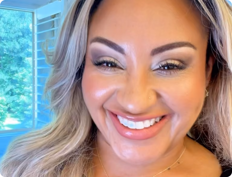 Close-up of a smiling woman with blonde hair and bright makeup beside a window with green outdoors.