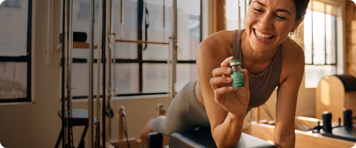 Smiling woman in workout clothes holding a small green vial labeled GLP-1 injection in gym setting.