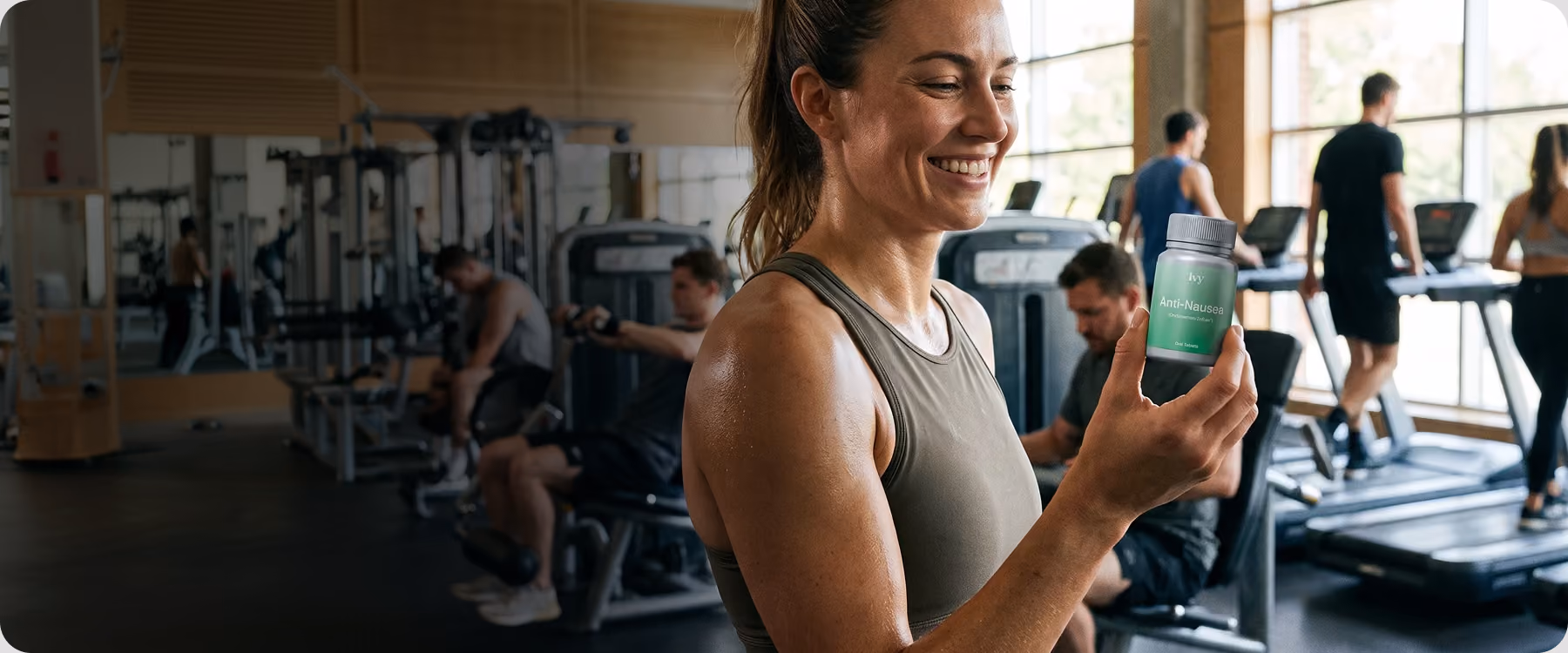 Smiling woman in gym holding a bottle of Ivy Anti-Nausea tablets with people exercising in background.