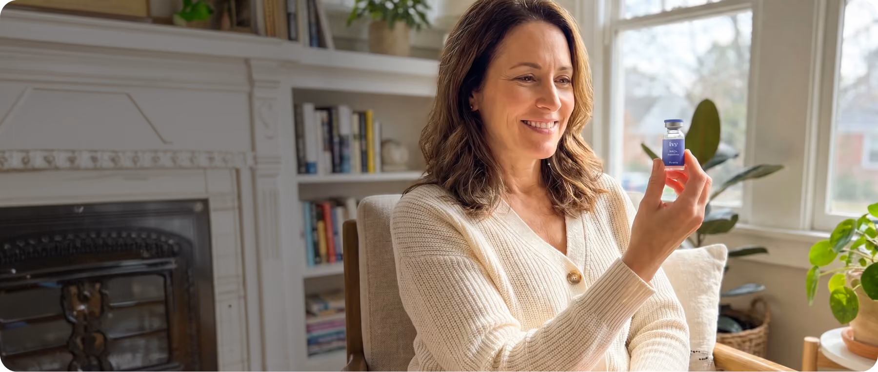 Smiling woman in a cream sweater holding a small bottle labeled Ivy NAD+ Injection by a window.