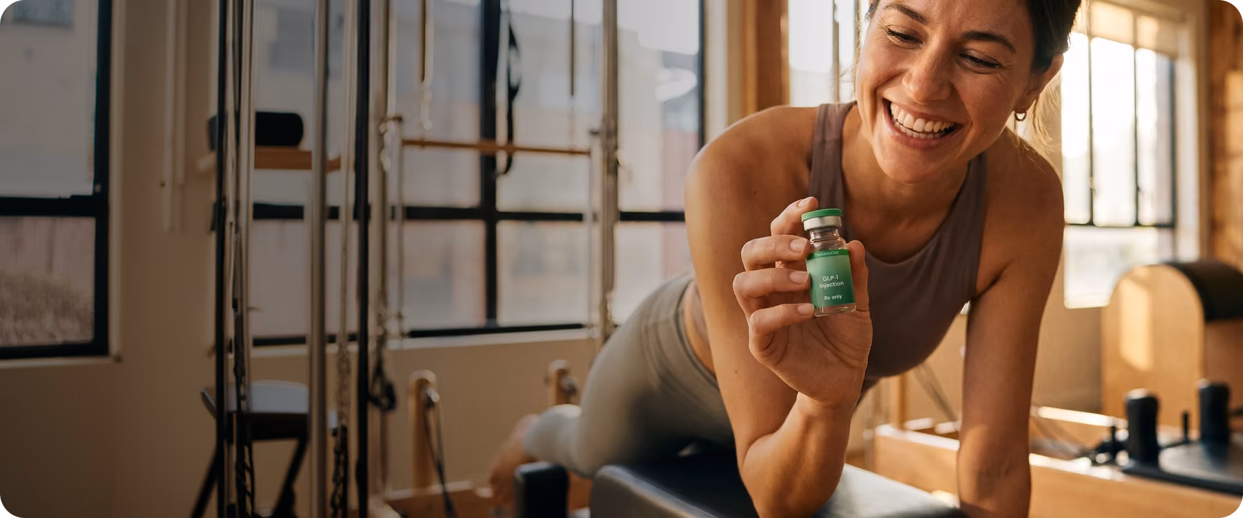Smiling woman in workout clothes holds a small green vial labeled GLP-1 injection in a fitness studio.